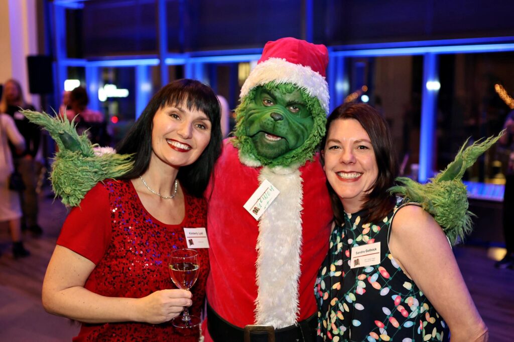 Women in festive attire posing with a character dressed as the Grinch at a holiday event, capturing a joyful moment of celebration and engagement.