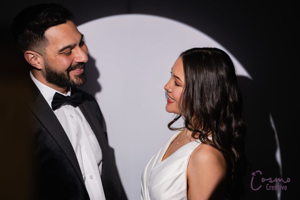 Elegant couple smiling in a dramatic photo booth setting, showcasing sophisticated style and memorable wedding experience, with soft lighting and a spotlight effect.