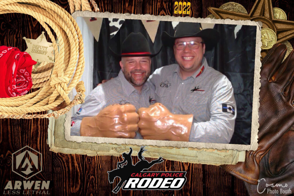 Two smiling men wearing cowboy hats and themed shirts, posing with oversized foam fists, framed by a decorative border featuring ropes and rodeo elements, celebrating the Calgary Police Rodeo event.
