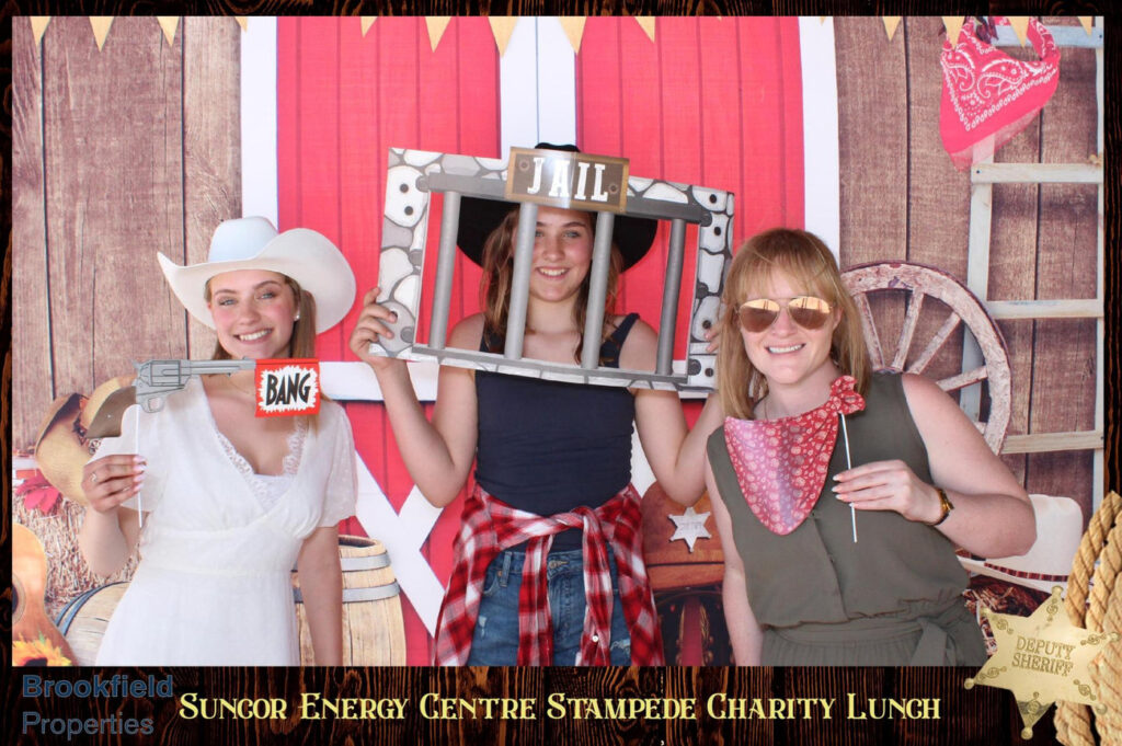 Three women posing in a western-themed photo booth at the Suncor Energy Charity Lunch during Calgary Stampede, featuring props like a toy gun and a jail frame, with festive decorations in the background.