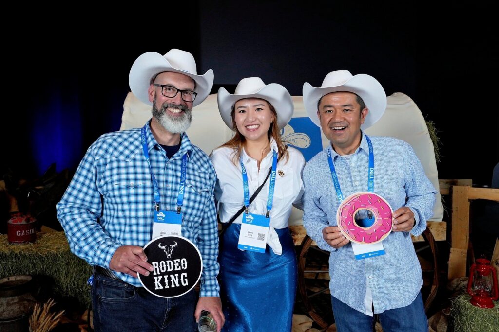Three individuals in cowboy hats posing at a Stampede-themed photo booth, holding props including a "Rodeo King" sign and a pink donut, with a festive backdrop and hay bales.