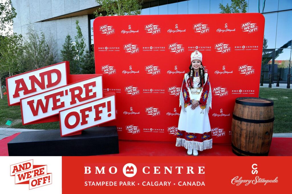 Woman in traditional attire standing in front of a red backdrop with "AND WE'RE OFF!" text, BMO Centre logo, and Calgary Stampede branding, celebrating the grand opening event in Calgary.