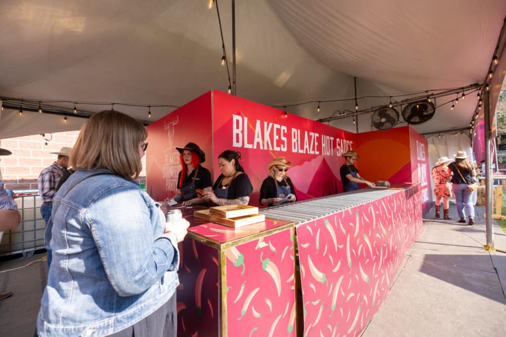 Blakes Blaze Hot Sauce booth at a corporate event, featuring staff serving guests, vibrant pink branding, and a lively outdoor setting.