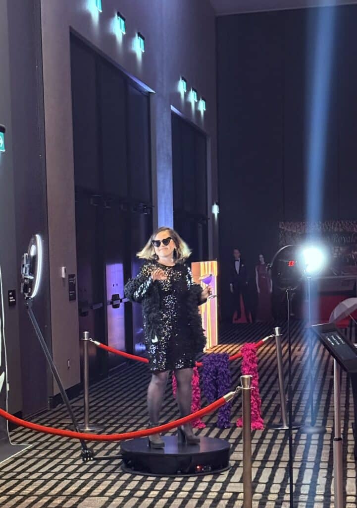 Woman in a sparkling black dress and sunglasses, engaging with guests at a 360 Video Booth, surrounded by red ropes and colorful decorations, creating an immersive event experience.