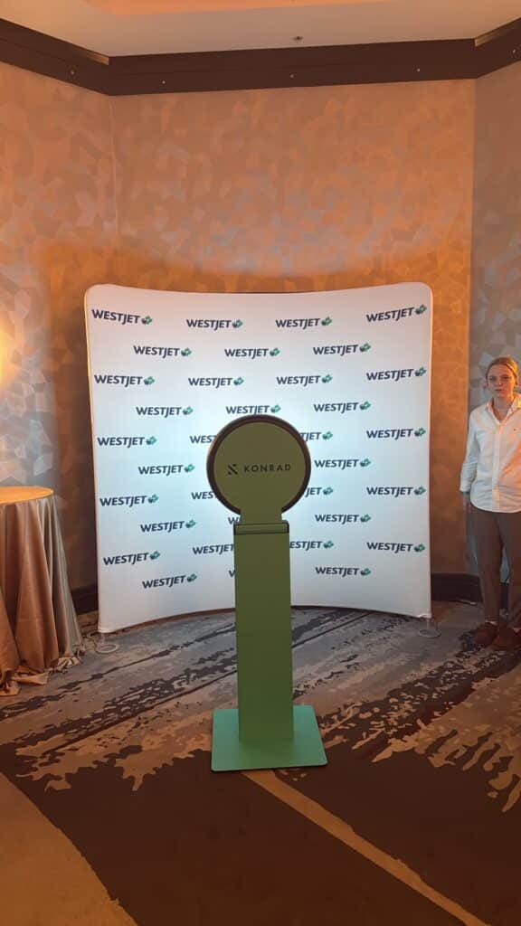 Digital photo booth setup featuring a green photo booth structure against a WestJet branded backdrop, with a woman in a white shirt standing nearby, ideal for event engagement and branding.