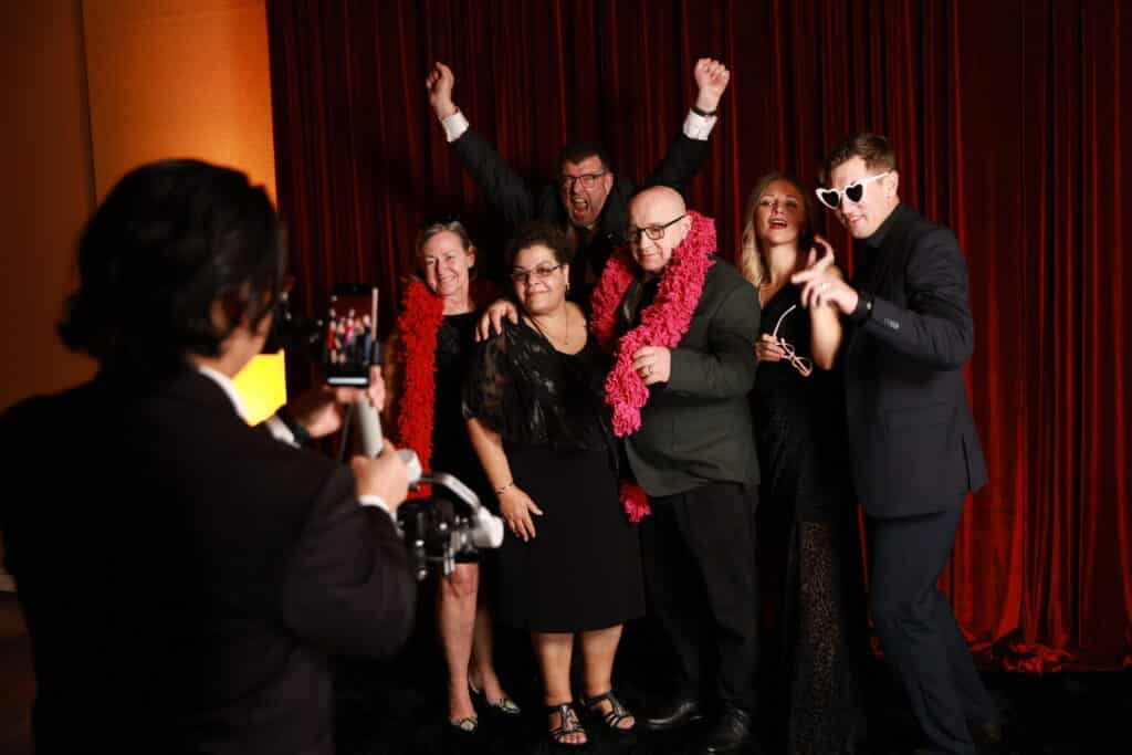 Group of six people posing joyfully in a photo booth with props, wearing festive attire, and surrounded by a vibrant red backdrop, capturing a lively moment perfect for event engagement and share-worthy content.