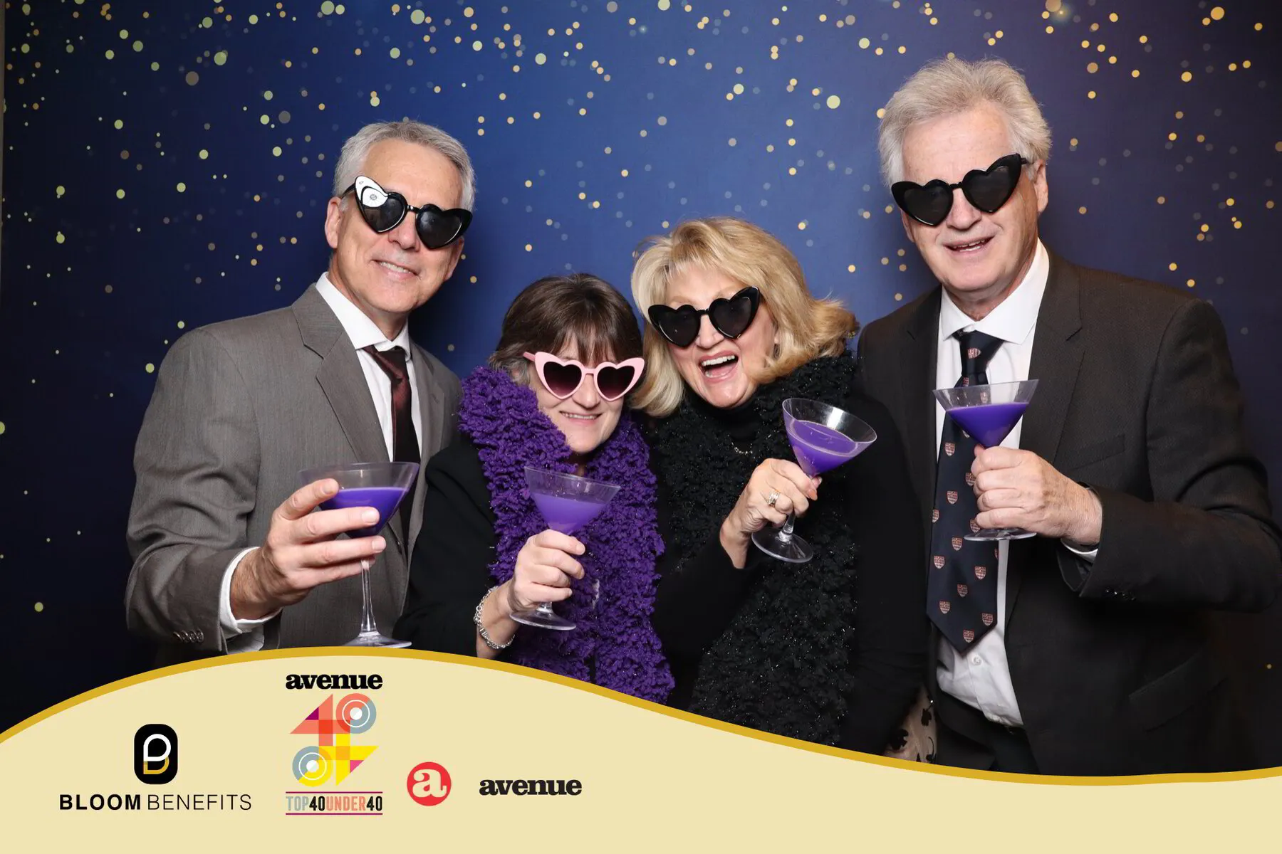 Group of four smiling individuals at the Avenue Magazine 40 Under 40 Awards, wearing heart-shaped sunglasses and holding purple cocktails, against a sparkling blue backdrop, featuring Bloom Benefits logo and event branding.