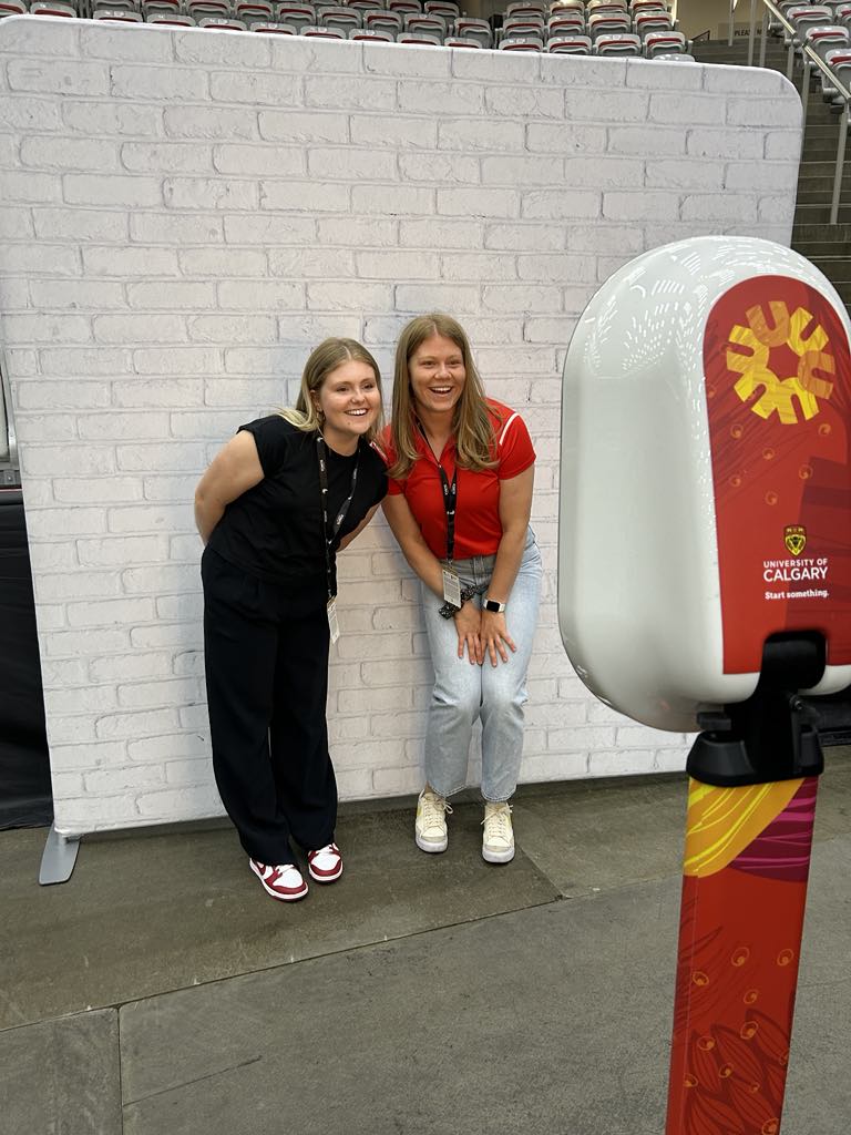 Two women posing playfully in front of a brick backdrop at a sports event, with a photo booth featuring the University of Calgary logo in the foreground, capturing the excitement of fan engagement and team spirit.