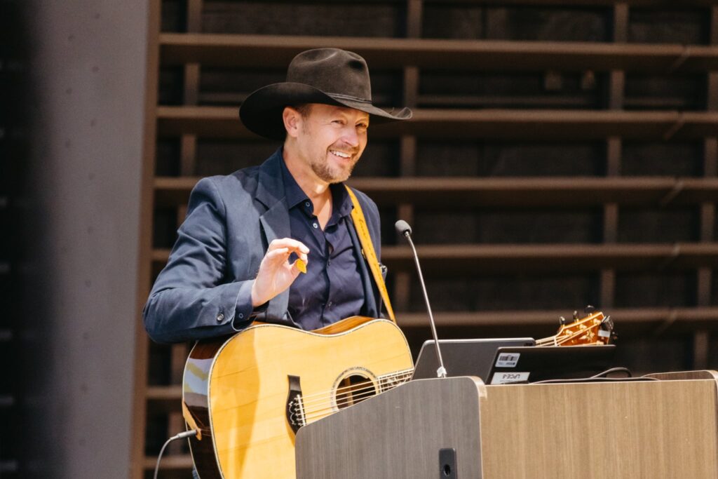 Man in a cowboy hat smiling while holding an acoustic guitar, engaging with an audience at a trade show presentation.