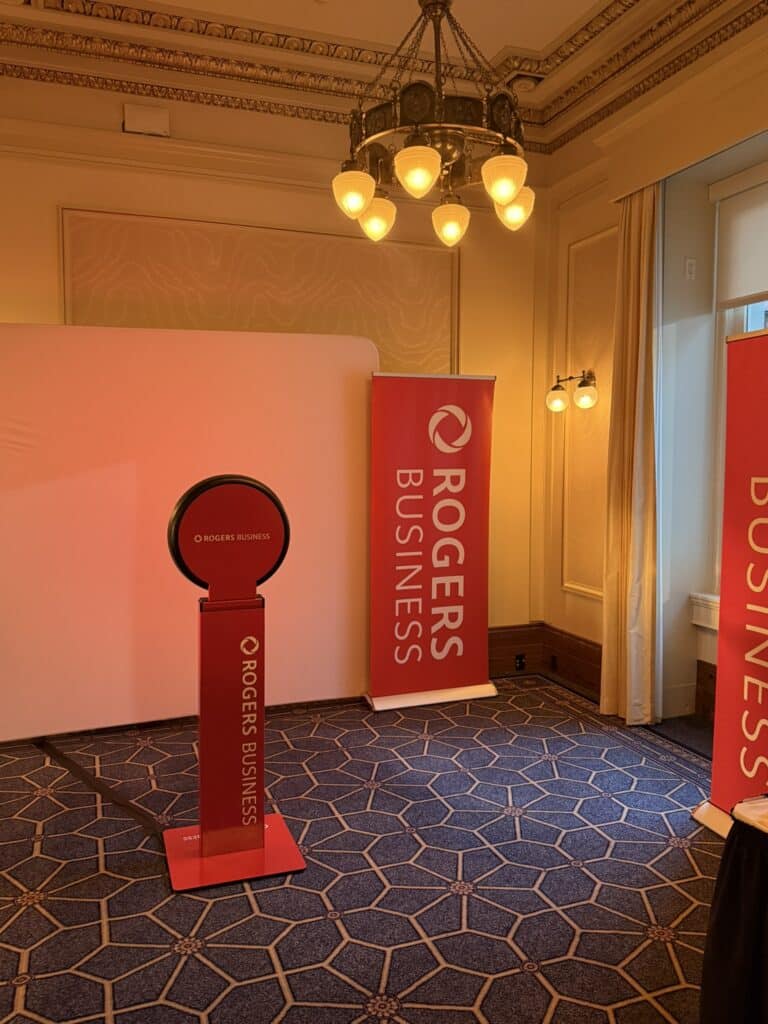 Branded photo booth setup featuring a circular camera stand and two red banners displaying "Rogers Business," set in an elegant event space with decorative lighting and patterned carpet.