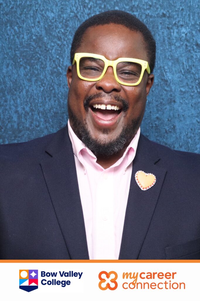 Professional headshot of a smiling man wearing yellow glasses and a blazer, with a pink shirt and a heart-shaped pin, set against a blue backdrop, featuring logos for Bow Valley College and My Career Connection.