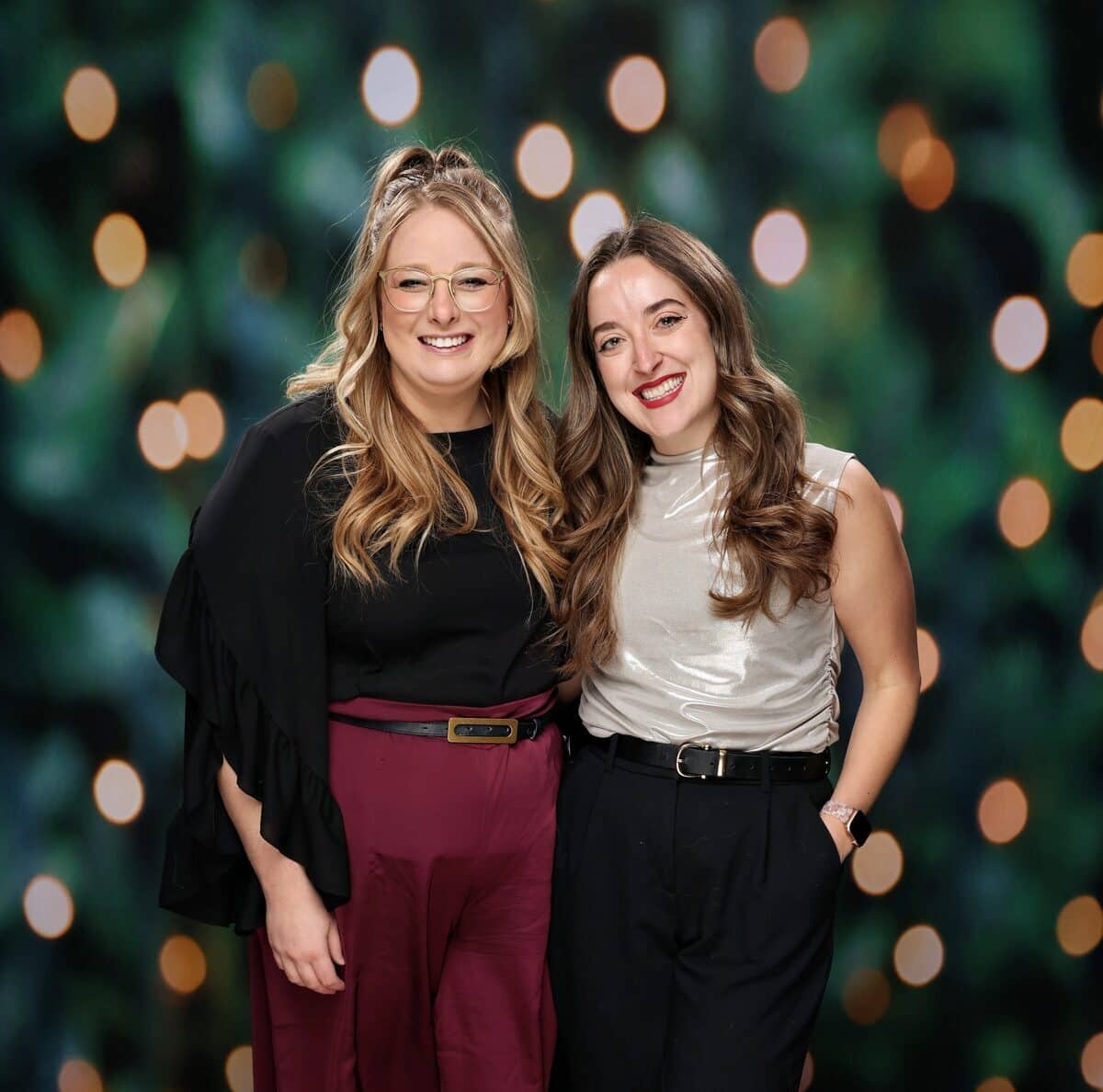 Two women posing together, one in a black top and burgundy pants, the other in a silver top and black pants, against a blurred green background with soft, glowing bokeh lights, representing an elegant photo moment for events.