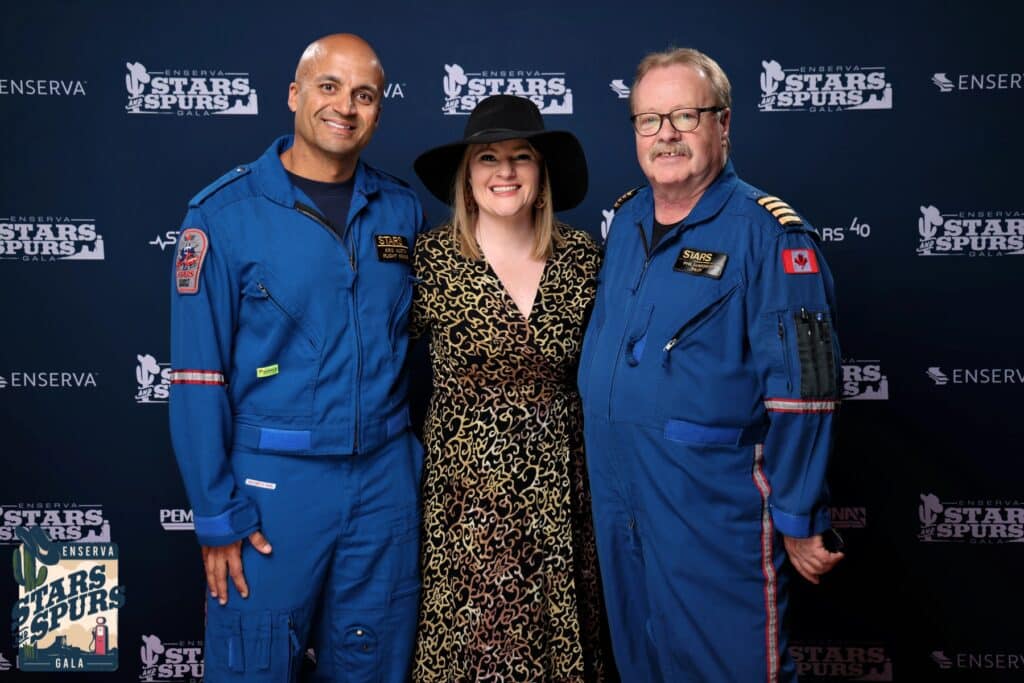 Group of three individuals posing for a photo at the Enserva Stars and Spurs Gala, featuring two men in blue flight suits and a woman in a patterned dress with a wide-brimmed hat, against a branded backdrop.