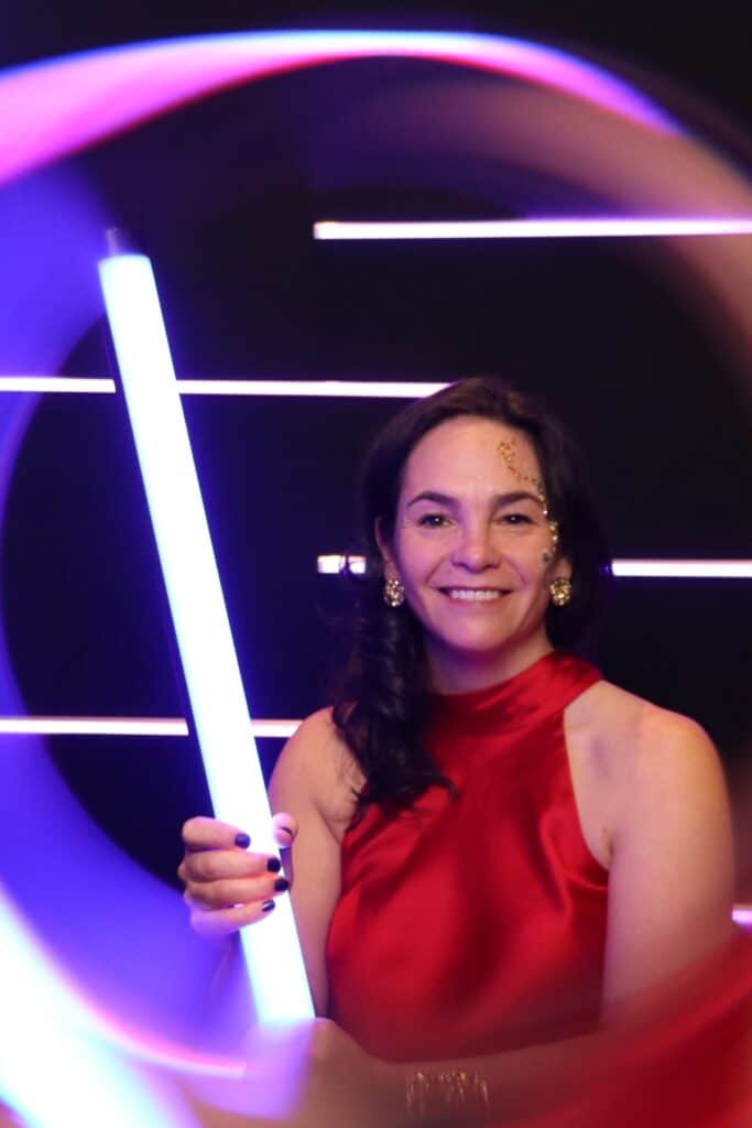 Woman in a red dress holding a glowing light stick, smiling against a vibrant backdrop with dynamic lighting, embodying the energetic atmosphere of the Glow Booth experience at events.