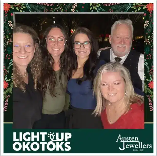 Group of five smiling individuals posing together at the Light Up Okotoks festival, featuring a branded border with "LIGHT UP OKOTOKS" and "Austen Jewellers" logos, capturing a festive moment during the event.
