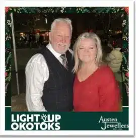 Couple posing together at the Light Up Okotoks Family Festival, featuring branded photo frame with "Light Up Okotoks" and "Austen Jewellers" logos, capturing festive event spirit.