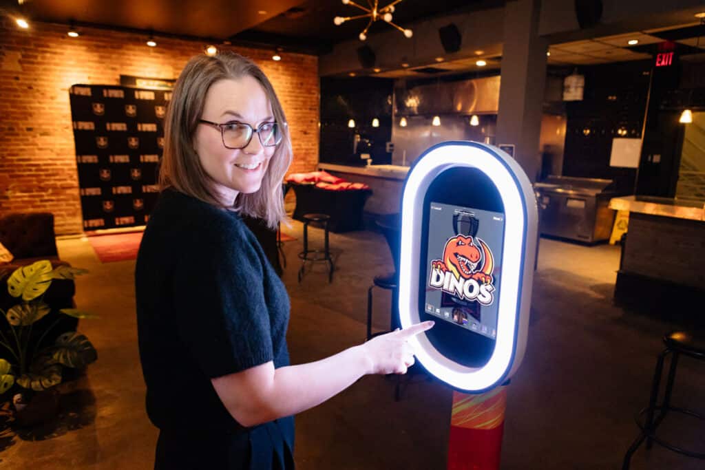 Woman interacting with a photo booth displaying a personalized trading card design featuring a dinosaur logo, in a stylish event space with brick walls and ambient lighting.