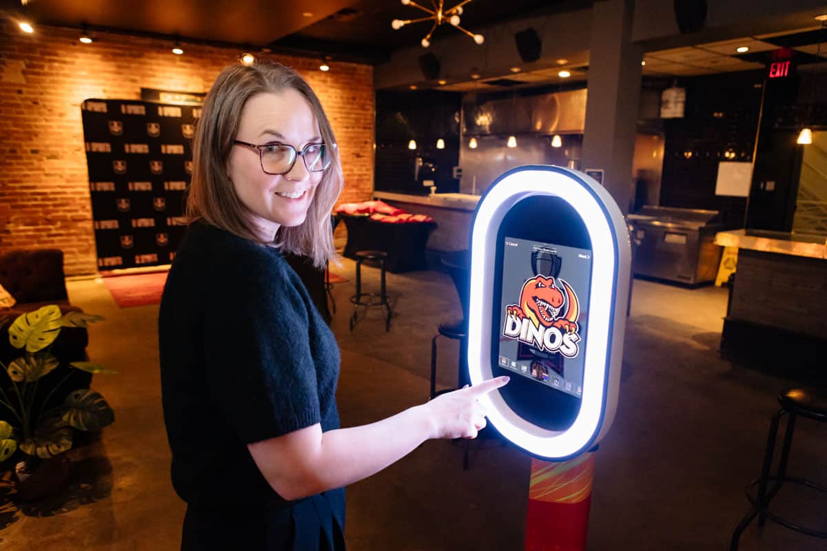 Woman interacting with a photo booth displaying a personalized trading card design featuring a dinosaur logo, in a stylish event space with brick walls and ambient lighting.