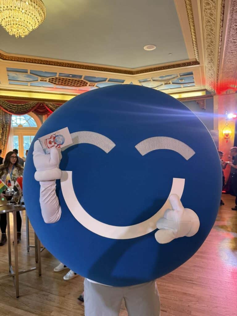 Blue mascot character with a smiling face holding a card, engaging attendees at a lively event, showcasing brand interaction and participant enjoyment.