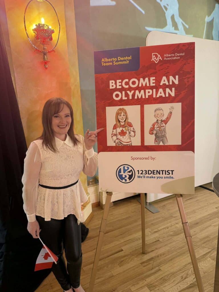 Woman smiling and posing next to a sign reading "Become an Olympian," sponsored by 123Dentist, at the Alberta Dental Team Summit event, featuring a Canadian flag and a vibrant background.
