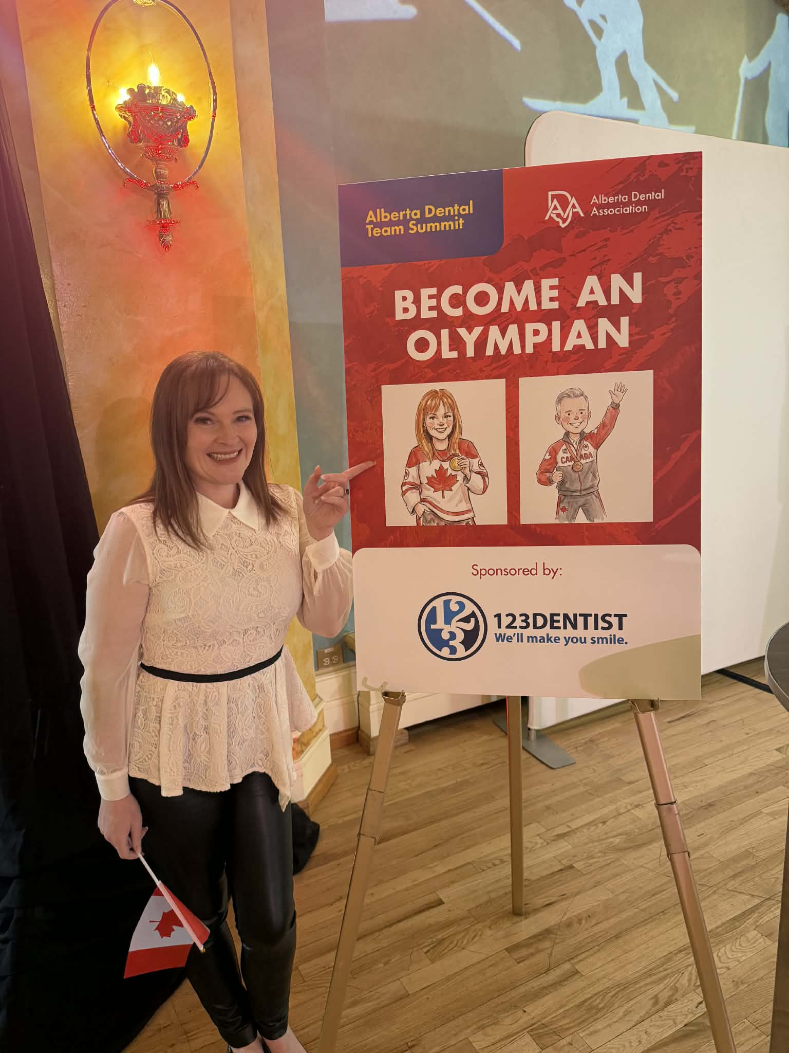 Woman smiling and posing next to a sign reading "Become an Olympian," sponsored by 123Dentist, at the Alberta Dental Team Summit event, featuring a Canadian flag and a vibrant background.