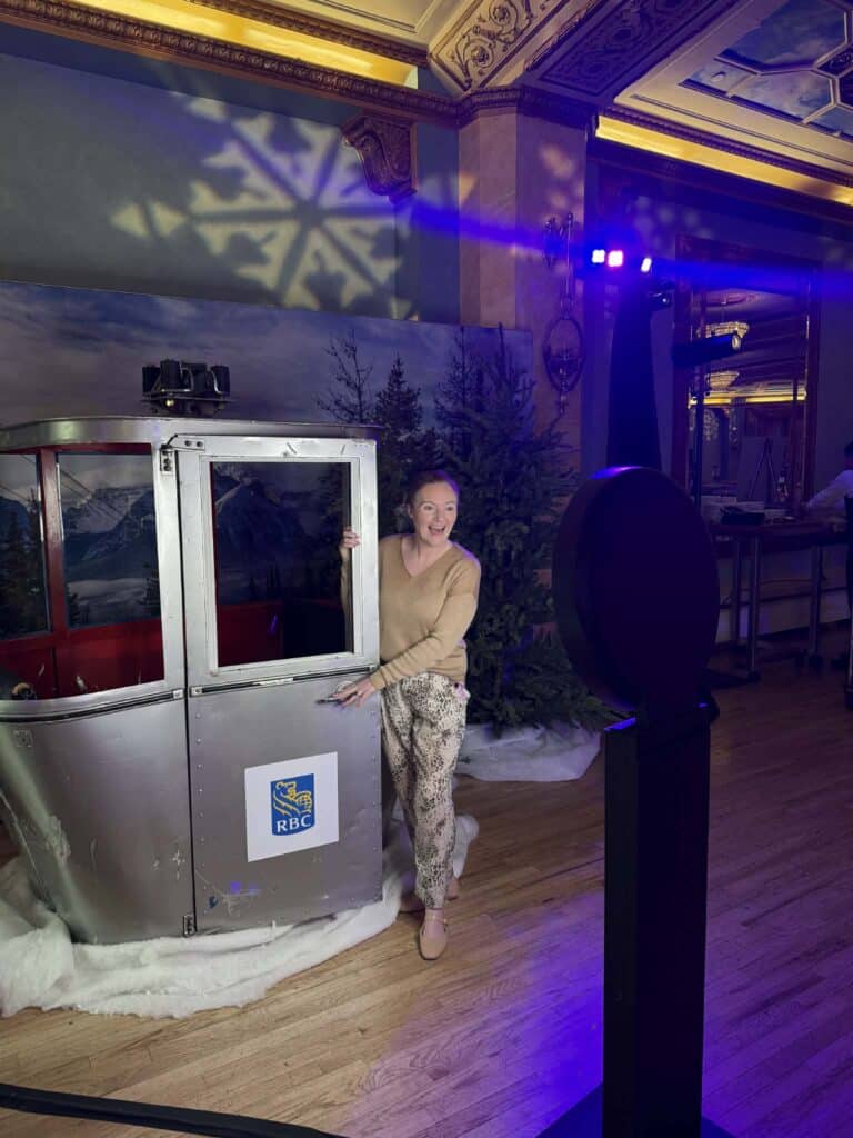 Woman posing beside an RBC-branded photo booth resembling a ski gondola, with festive decorations and lighting, at the Alberta Dental Conference mixer event.