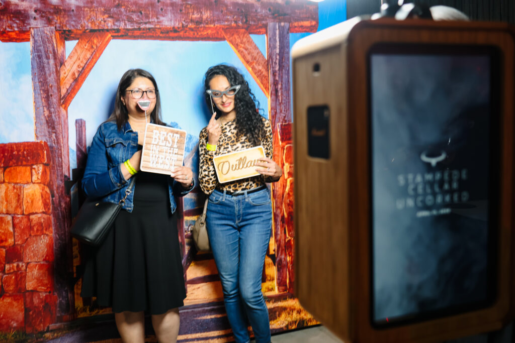 Two women posing in front of a rustic backdrop at the Stampede Cellar Uncorked event, holding playful signs that read "BEST IN THE WEST" and "Ouilian," with a photo booth in the foreground featuring the event logo.