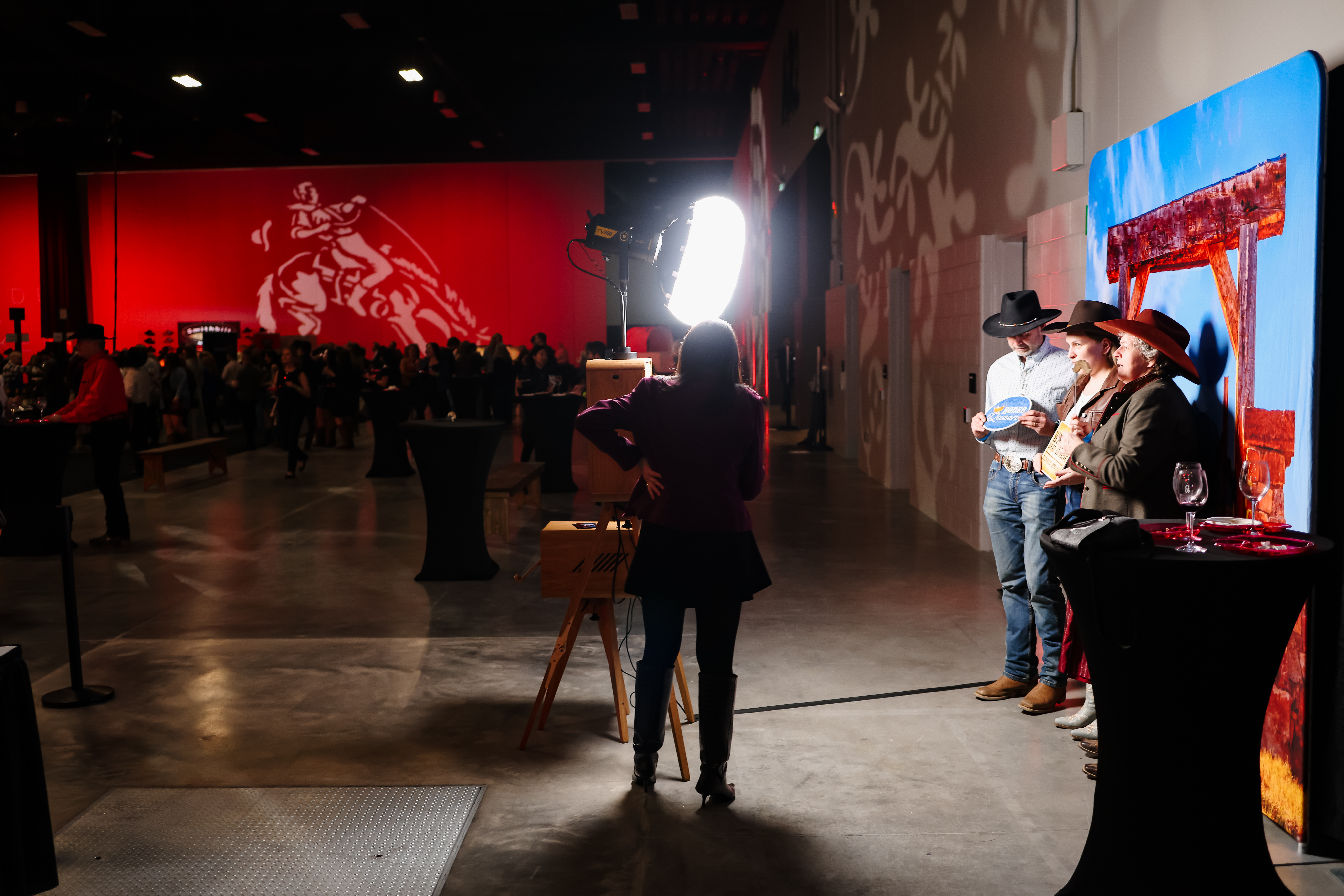 Guests enjoying a photo booth experience at the Stampede Cellar Uncorked event, featuring a vibrant red backdrop and western-themed props, with one guest holding a custom label while others pose in cowboy hats.