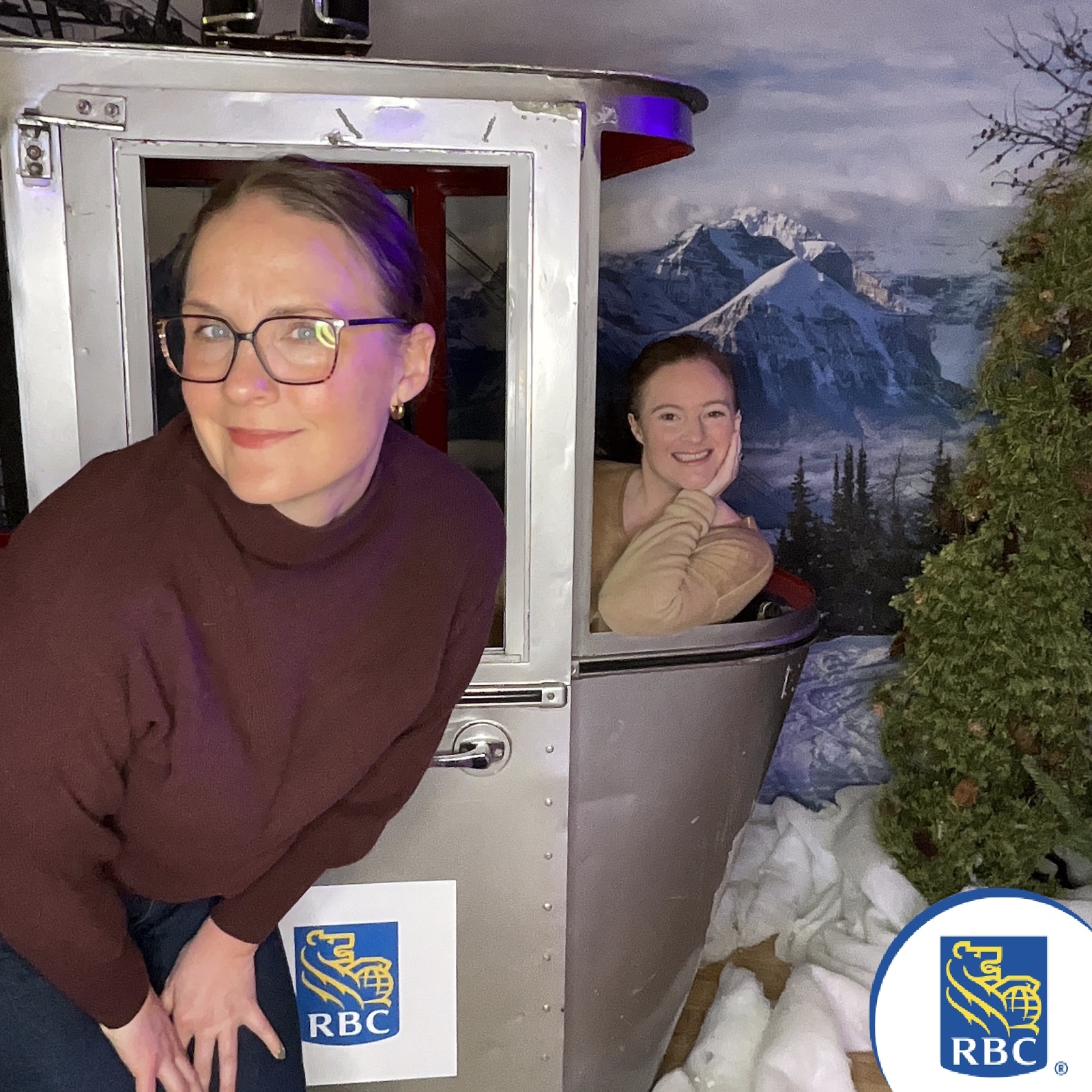 Two women posing in a photo booth designed like a ski gondola, showcasing RBC branding, with a mountain backdrop and winter-themed decor.
