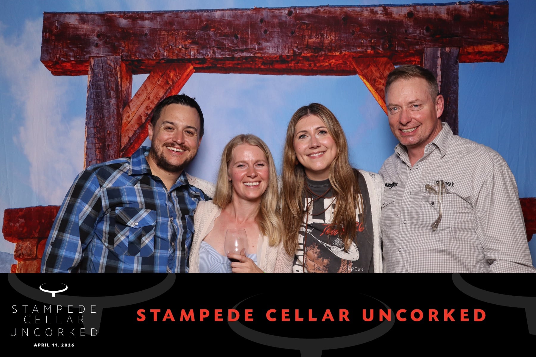 Group of four smiling guests at the Stampede Cellar Uncorked event, posing in front of a rustic wooden backdrop, holding wine glasses and enjoying the interactive photo booth experience.