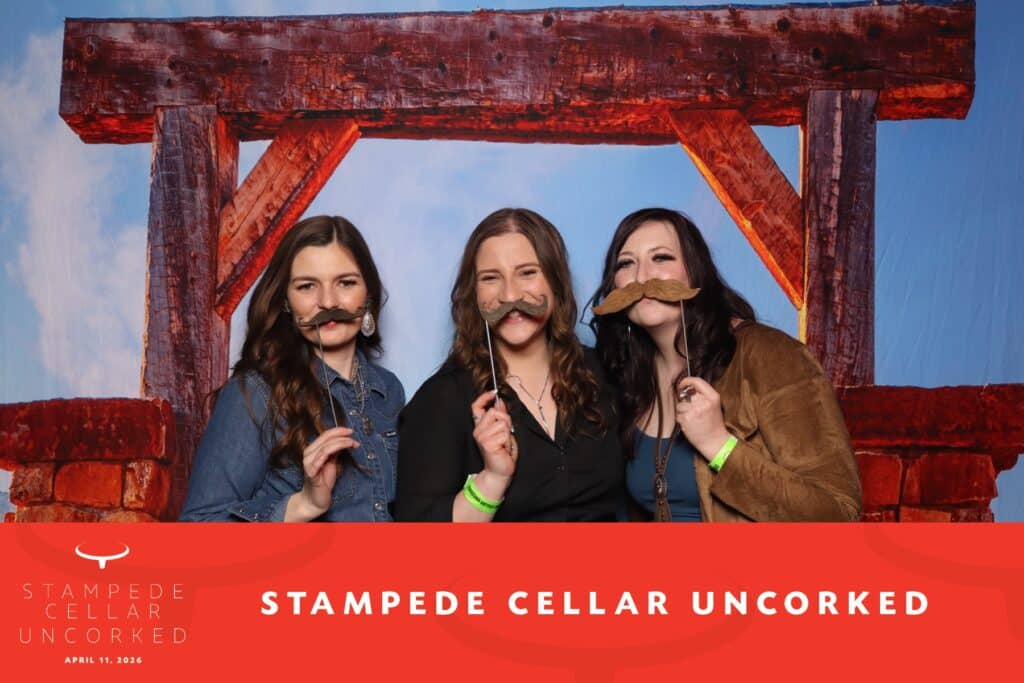 Three women posing with playful mustaches in front of a rustic backdrop at the Stampede Cellar Uncorked event, featuring a bold graphic with the event name and date.