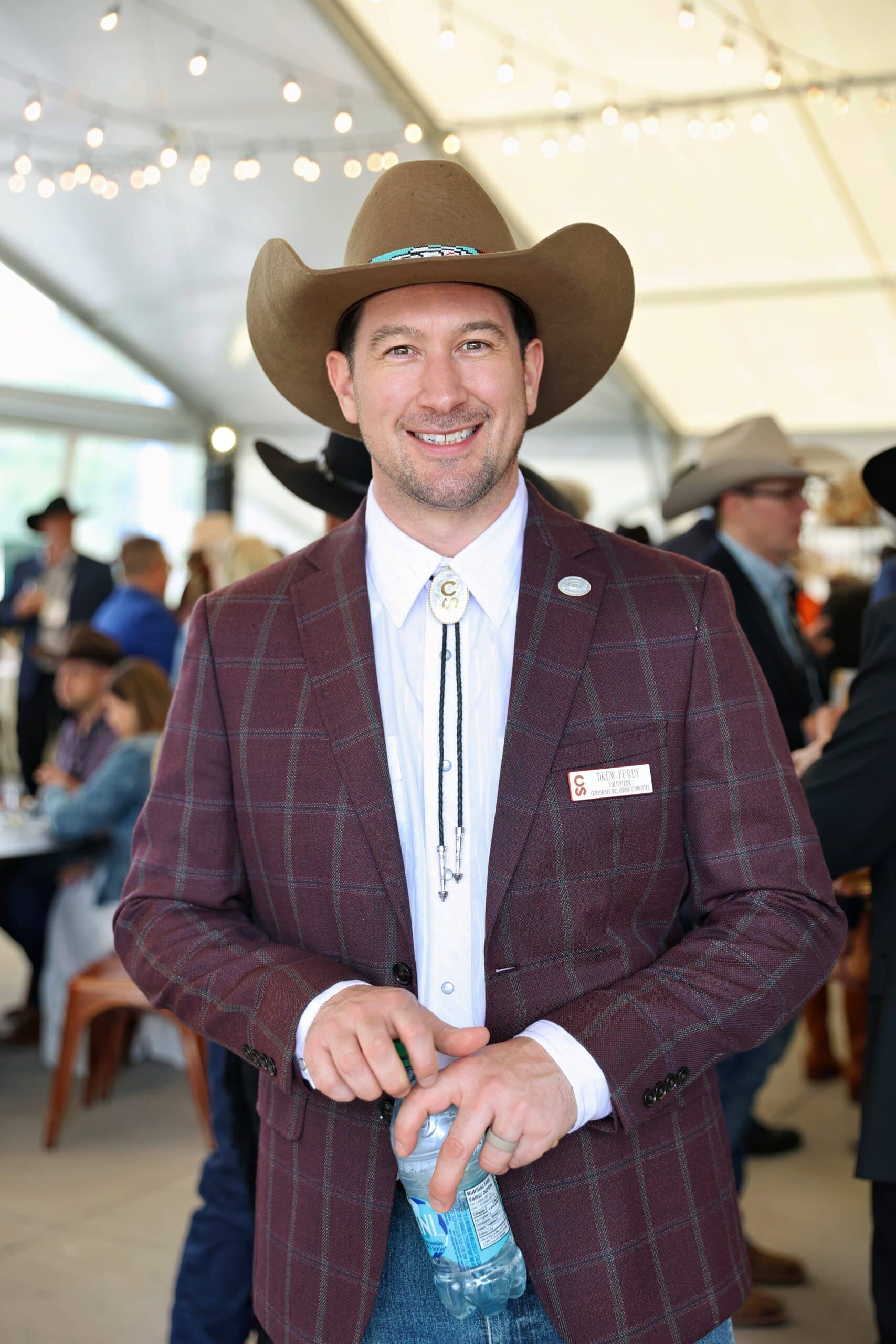 Man wearing a cowboy hat and a plaid blazer, smiling and holding a water bottle